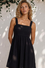 Woman wearing a black dress standing against a white wall with greenery.