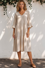 Woman wearing a beige dress standing against a white wall with greenery.