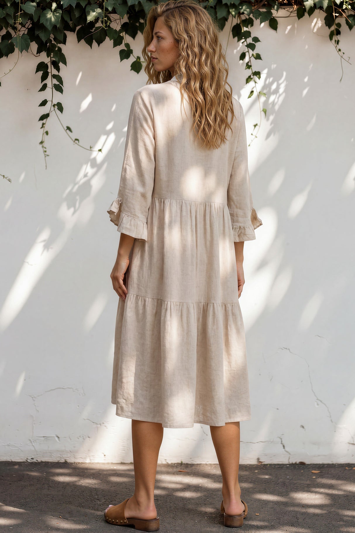 Woman wearing a beige dress standing against a white wall with greenery.