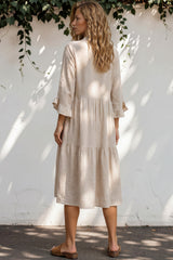 Woman wearing a beige dress standing against a white wall with greenery.