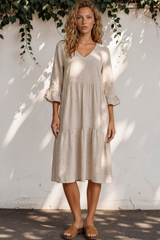 Woman wearing a beige dress standing against a white wall with greenery.