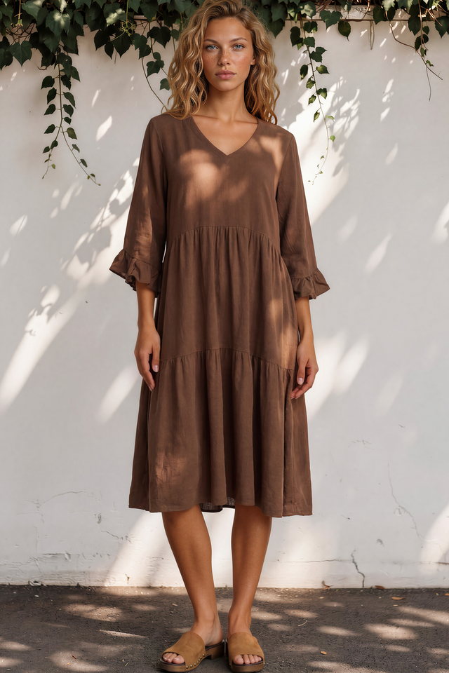 Woman wearing a brown dress standing against a white wall with greenery.