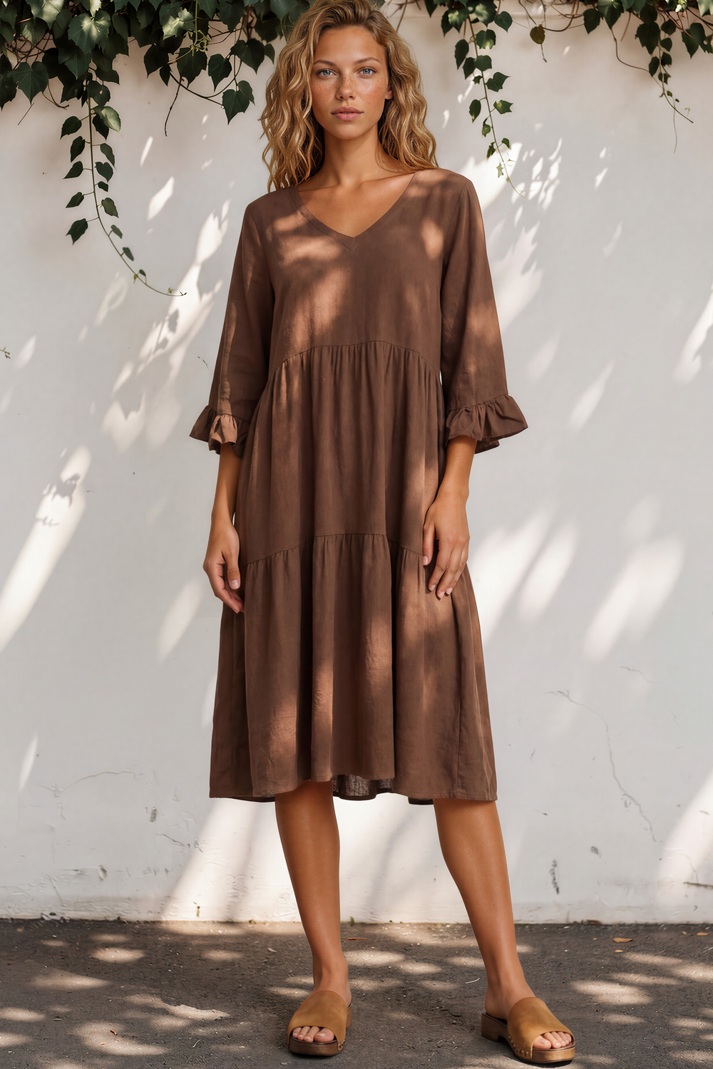 Woman wearing a brown dress standing against a white wall with greenery.