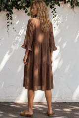 Woman wearing a brown dress standing against a white wall with greenery.