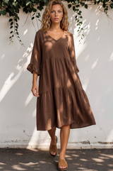 Woman wearing a brown dress standing against a white wall with greenery.