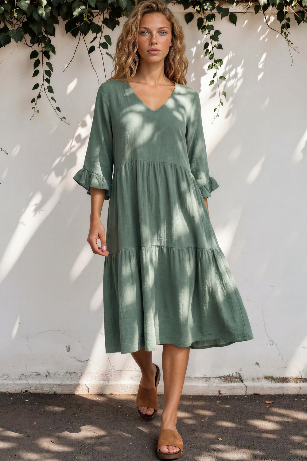 Woman wearing a green dress standing against a white wall with shadows and plants.