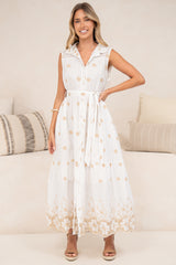 A woman wearing the Bruna Midi Dress in Palmira Print with one hand on her hip, showing the white collared button-front bodice, tie waist, peach floral embroidery and lace-trimmed hem, styled with tan sandals in front of a cream sofa