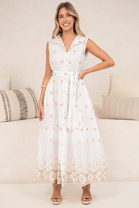 A woman wearing the Bruna Midi Dress in Palmira Print with one hand on her hip, showing the white collared button-front bodice, tie waist, peach floral embroidery and lace-trimmed hem, styled with tan sandals in front of a cream sofa