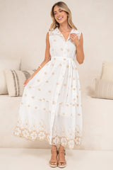 A smiling woman holding out the skirt of the Bruna Midi Dress in Palmira Print to show its full white midi shape, peach floral embroidery and lace-trimmed hem, with a collared sleeveless bodice and tan sandals in front of a cream sofa
