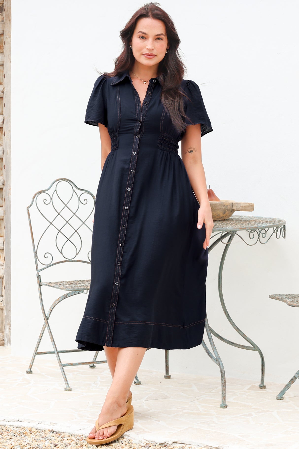 A woman modeling a dark navy midi shirt dress with short puff sleeves, a button-down front, and a gathered waist, standing in front of a white wall with a metal garden table and chair.