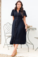 A woman modeling a dark navy midi shirt dress with short puff sleeves, a button-down front, and a gathered waist, standing in front of a white wall with a metal garden table and chair.