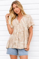 A woman wearing a beige short-sleeve button-up blouse with a white leaf pattern and light blue denim shorts, standing against a white paneled wall.