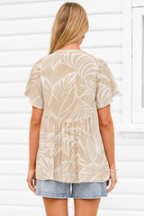 A woman viewed from behind wearing a beige short-sleeve top with a white tropical leaf pattern, paired with denim shorts, standing against a white paneled wall.
