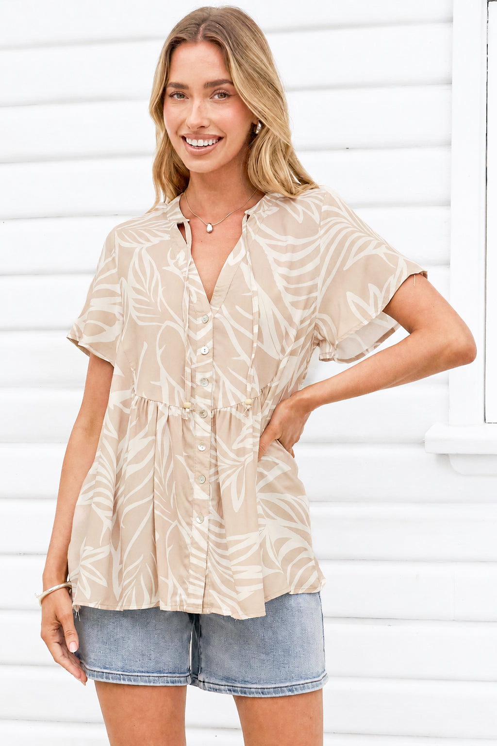 A woman wearing a beige short-sleeve button-front top with a white tropical leaf print, paired with denim shorts, standing against a white siding background.