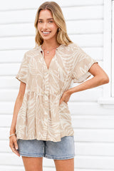 A woman wearing a beige short-sleeve button-front top with a white tropical leaf print, paired with denim shorts, standing against a white siding background.