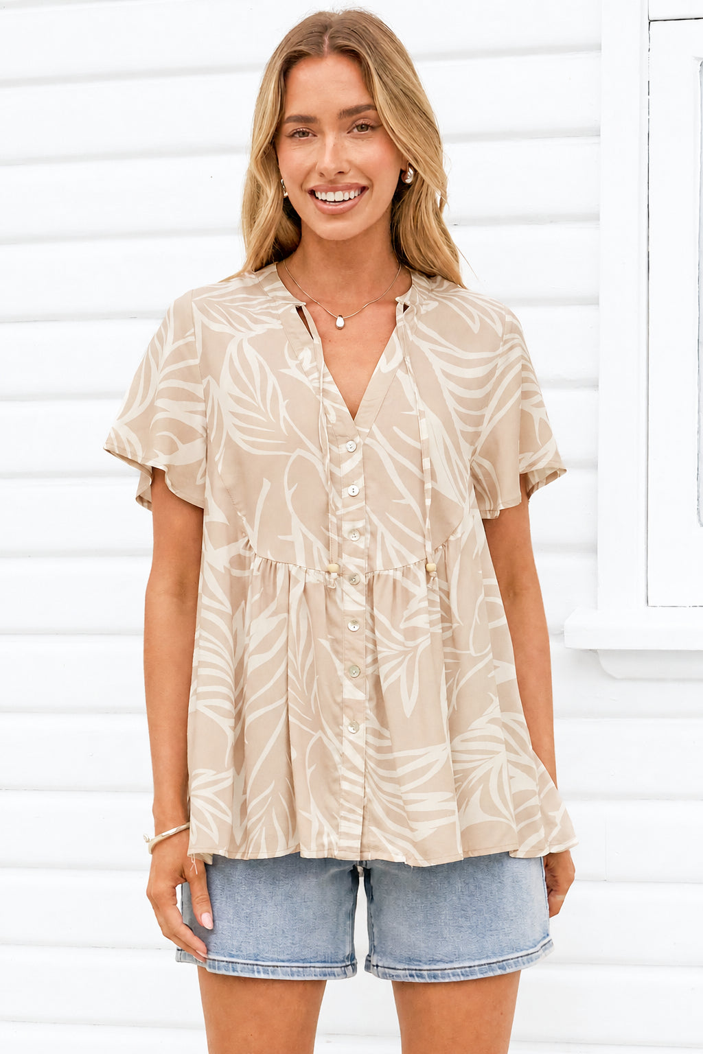 A woman wearing a beige short-sleeve button-front blouse with a white tropical leaf pattern, paired with denim shorts, standing in front of a white wooden wall.
