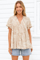 A woman wearing a beige short-sleeve button-front blouse with a white tropical leaf pattern, paired with denim shorts, standing in front of a white wooden wall.