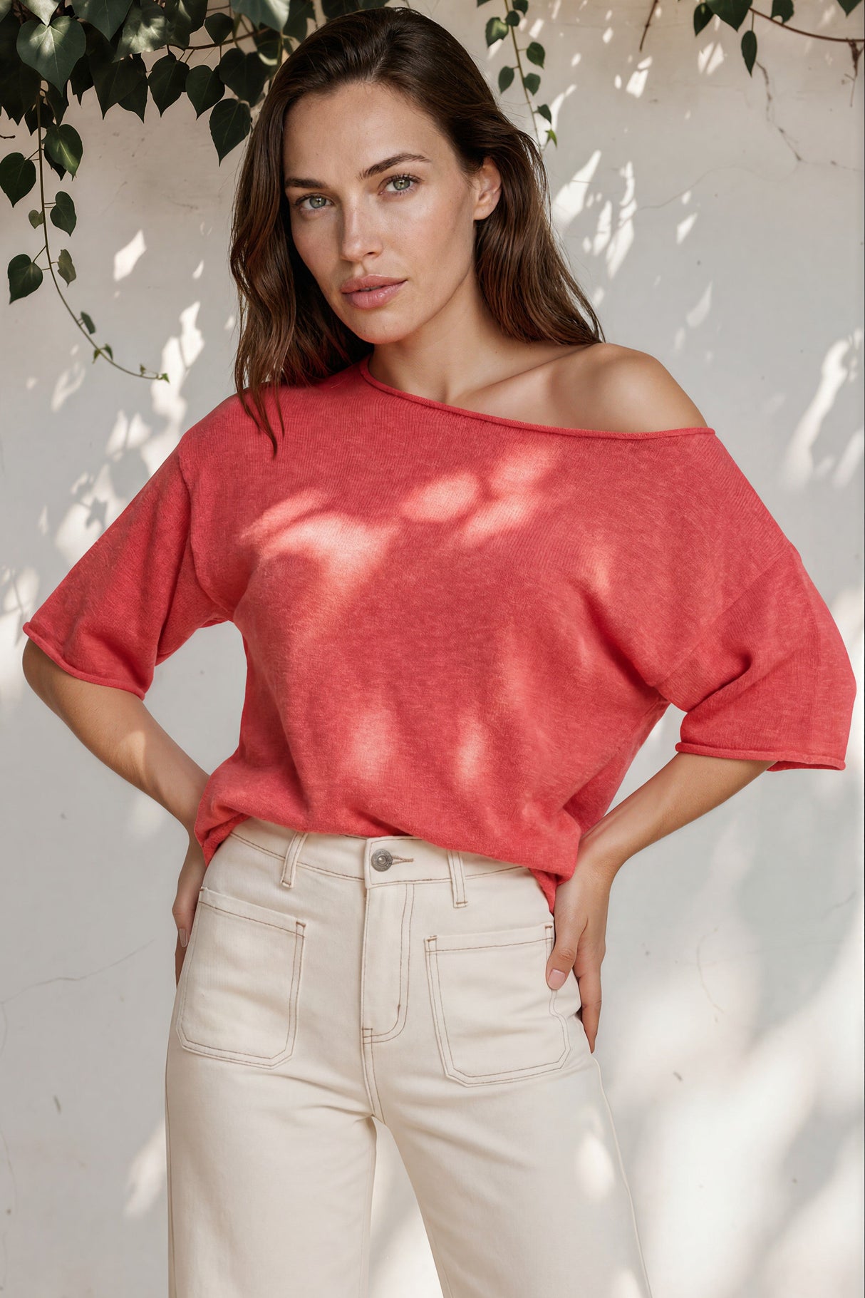 Woman wearing a red off-shoulder top and white pants standing against a white wall with greenery.