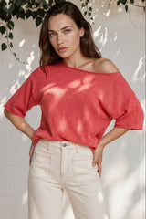 Woman wearing a red off-shoulder top and white pants standing against a white wall with greenery.