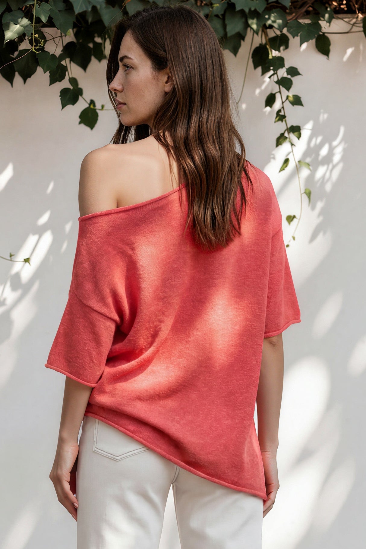 Woman wearing a coral off-shoulder top and white pants standing against a white wall with greenery.