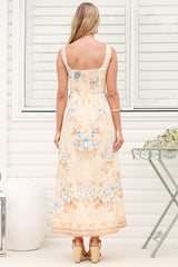 A woman wearing the Camelia midi dress in a dusty blue and tan floral print, featuring a square neckline and wide straps, standing in front of a white panelled wall.