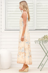 A woman wearing the Camelia midi dress in a dusty blue and tan floral print, featuring a square neckline and wide straps, standing in front of a white panelled wall.