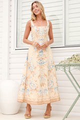 A woman wearing the Camelia midi dress in a dusty blue and tan floral print, featuring a square neckline and wide straps, standing in front of a white panelled wall.