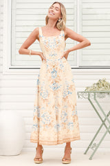 A woman wearing the Camelia midi dress in a dusty blue and tan floral print, featuring a square neckline and wide straps, standing in front of a white panelled wall.