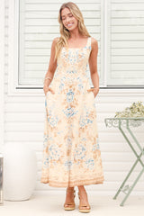 A woman wearing the Camelia midi dress in a dusty blue and tan floral print, featuring a square neckline and wide straps, standing in front of a white panelled wall.