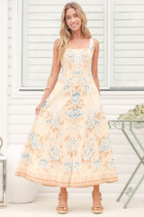 A woman wearing the Camelia midi dress in a dusty blue and tan floral print, featuring a square neckline and wide straps, standing in front of a white panelled wall.