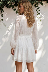 Woman wearing a white dress standing against a white wall with greenery.