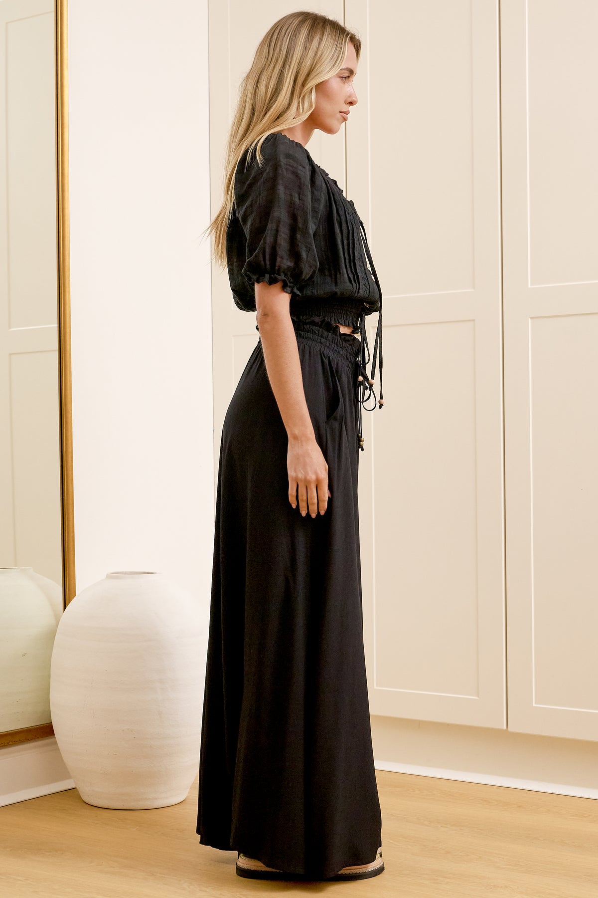 Wearing high-waisted black pants with a gathered paper-bag waist and wide-leg fit, a woman stands in front of a warm beige closet.