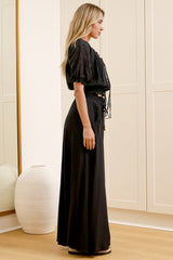 Wearing high-waisted black pants with a gathered paper-bag waist and wide-leg fit, a woman stands in front of a warm beige closet.
