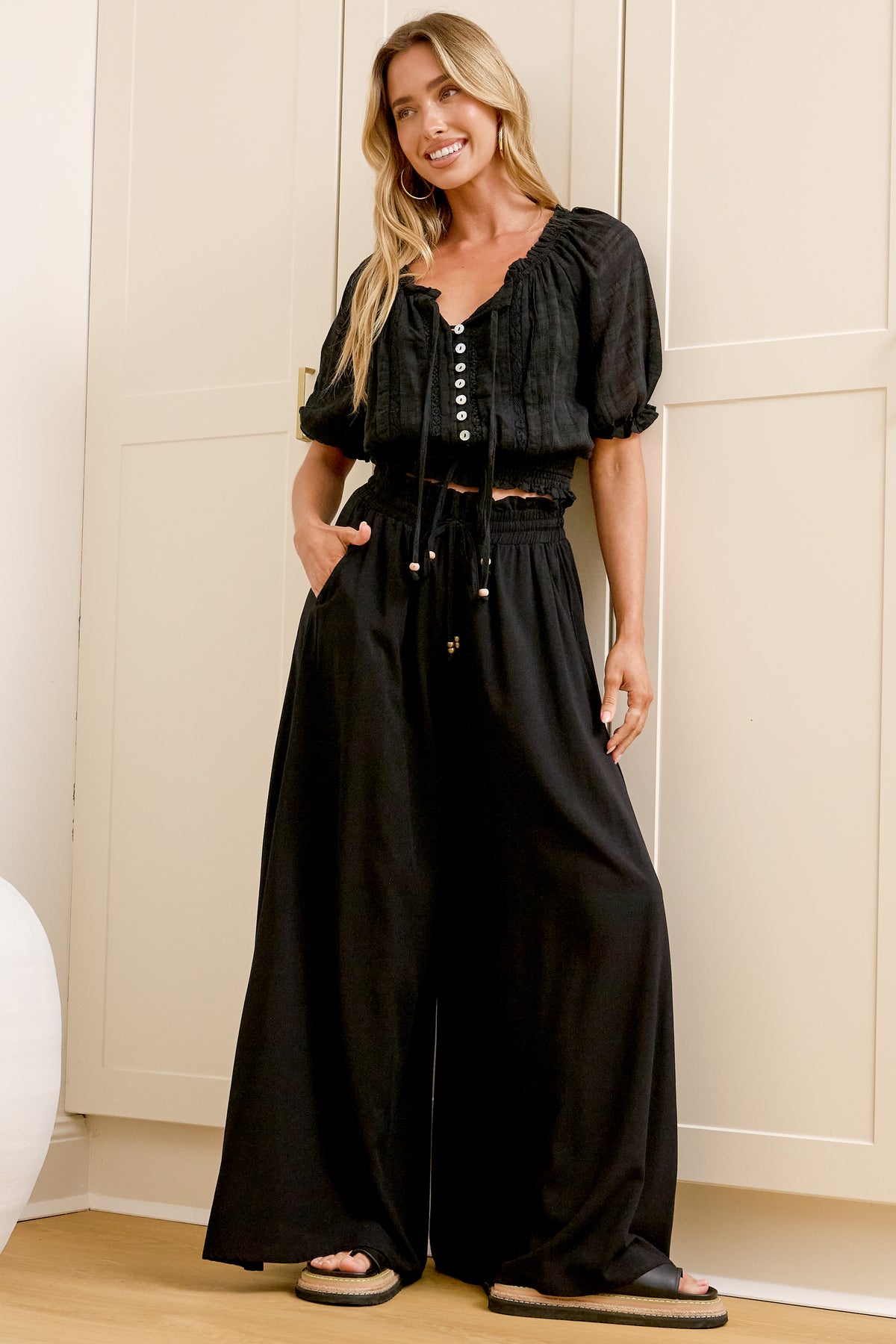 Wearing high-waisted black pants with a gathered paper-bag waist and wide-leg fit, a woman stands in front of a warm beige closet.