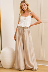 A woman wearing sand-coloured wide-leg pants with a high elasticated waist and relaxed fit, standing in front of a warm beige closet.