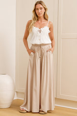 A woman wearing sand-coloured wide-leg pants with a high elasticated waist and relaxed fit, standing in front of a warm beige closet.