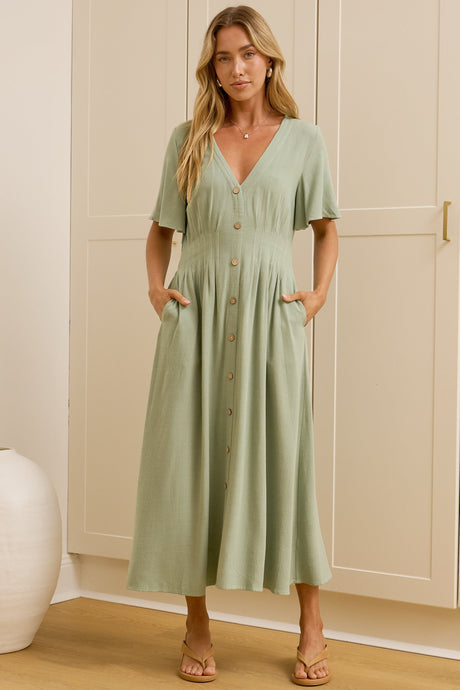 A woman wearing a sage midi dress with a V-neckline, flutter sleeves, and shirred elastic waistband, standing in front of a warm beige closet.