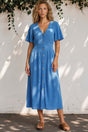 Woman wearing a blue dress standing against a white wall with greenery.