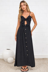A woman modeling a long black maxi dress with a sweetheart neckline, front keyhole cutout, and a row of gold buttons down the front, standing in a minimalist room with a white brick wall and beige rug. 