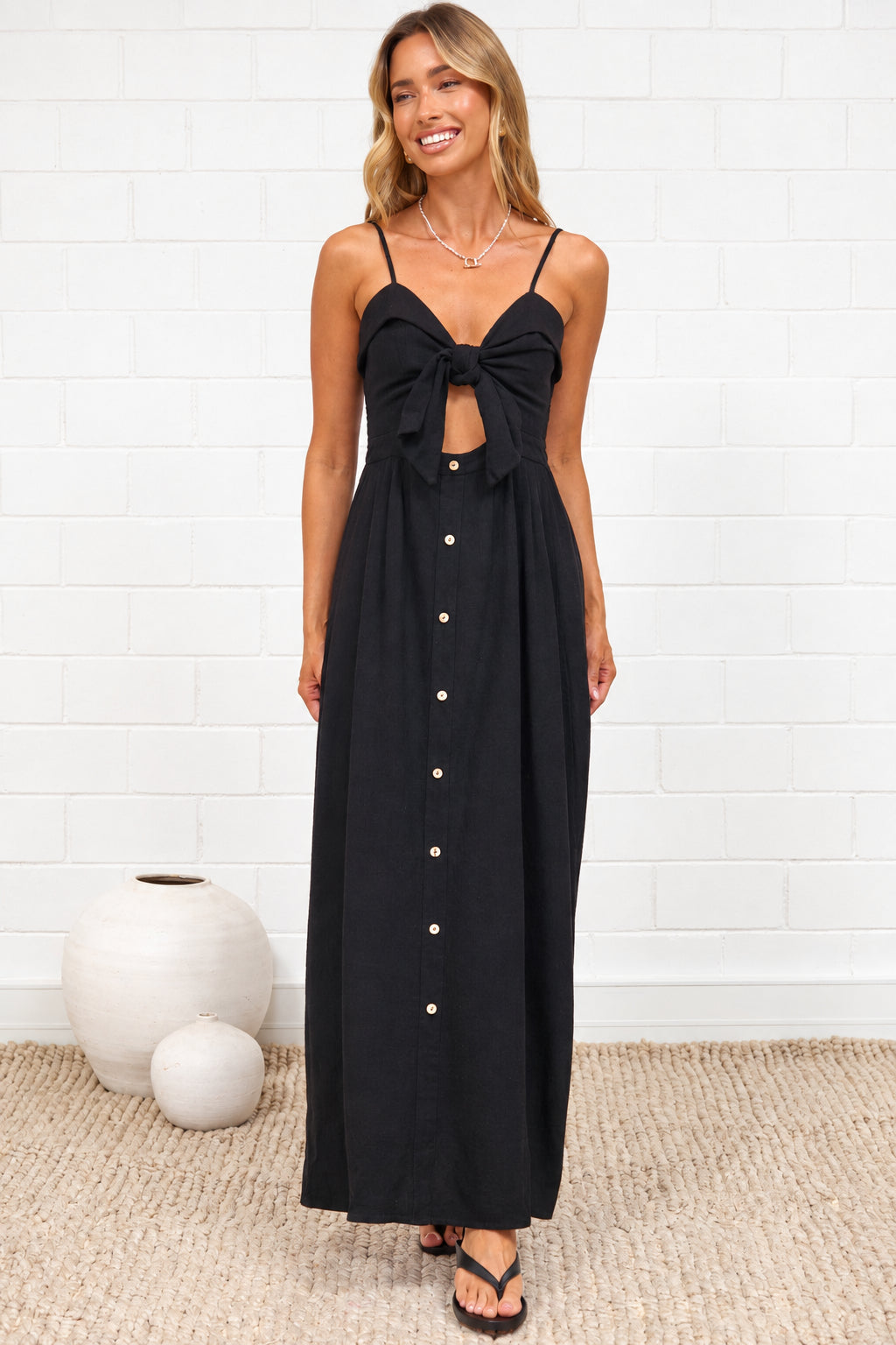 A woman wearing a black midi dress with a tie-front neckline, cutout detail, adjustable spaghetti straps, and decorative button placket, standing against a white painted brick wall.