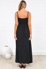 A woman wearing a black midi dress with a tie-front neckline, cutout detail, adjustable spaghetti straps, and decorative button placket, standing against a white painted brick wall.
