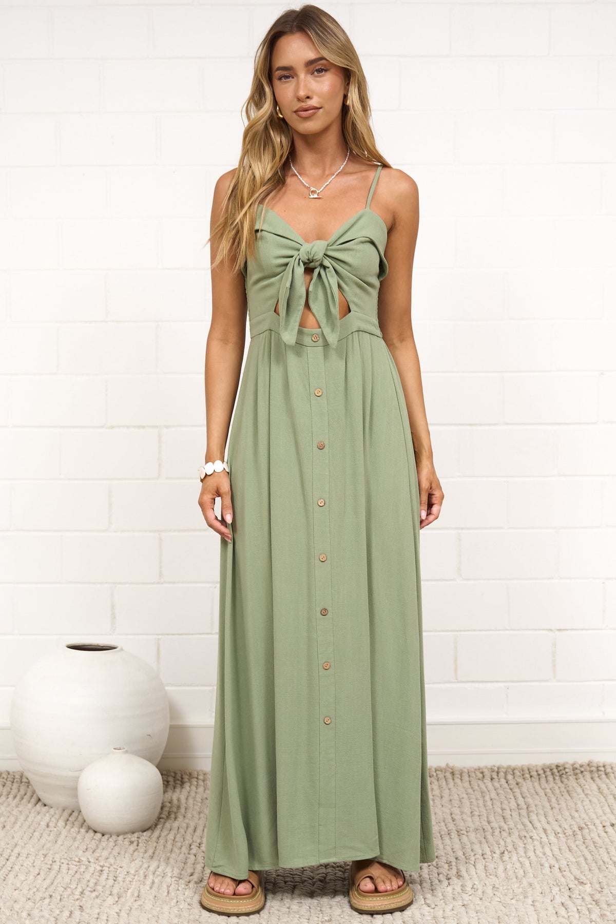 A woman modeling a sage green maxi dress with a tie-front detail and button-down front, standing on a woven rug against a white brick wall. 