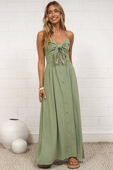 A woman stands in a bright room wearing a long sage green maxi dress with a tie-neck detail and button front. The room has a white brick wall, a beige rug, and white decorative vases. 