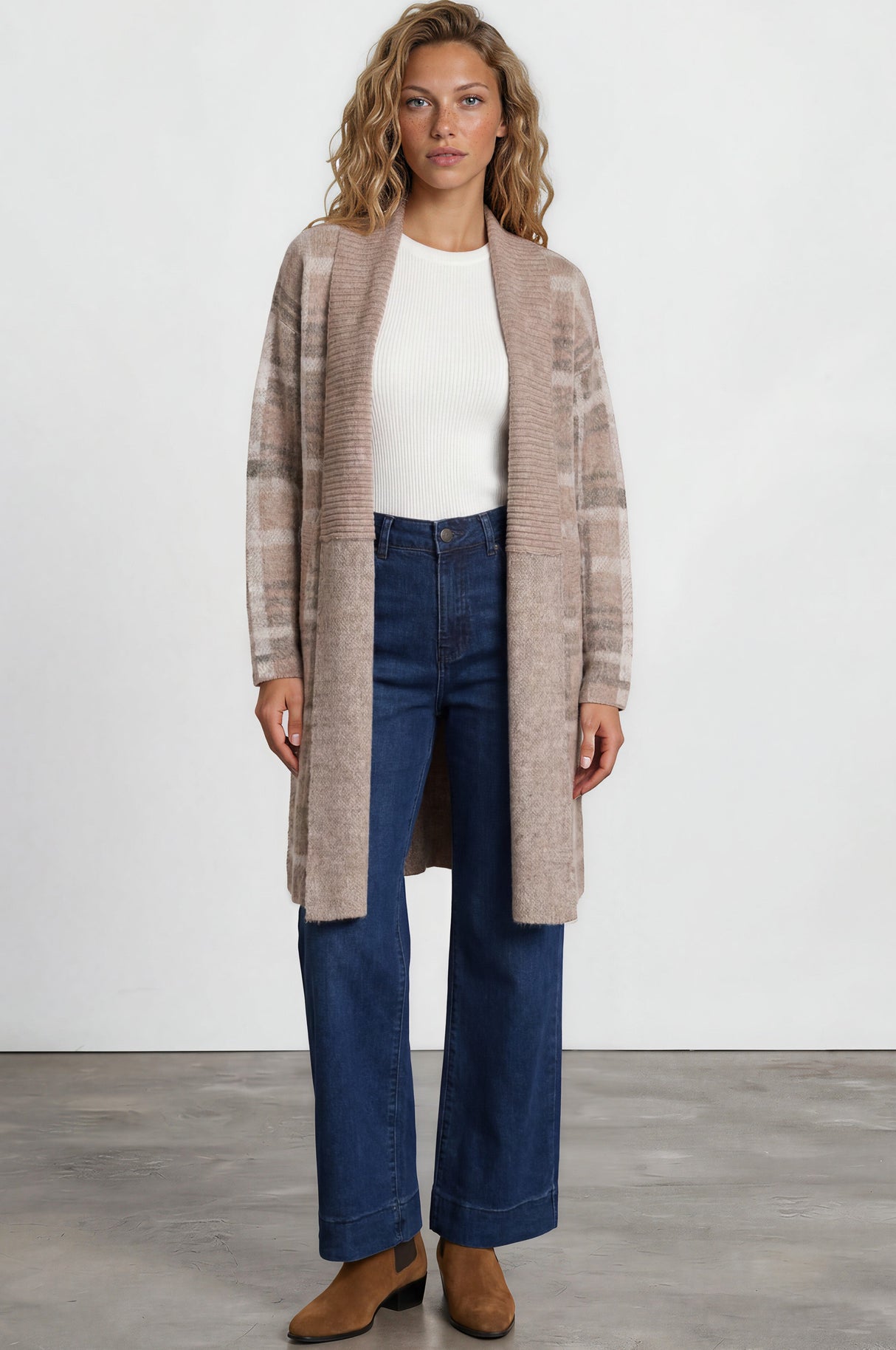 Woman wearing a long beige cardigan, white top, blue jeans, and brown shoes on a white background