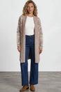 Woman wearing a long beige cardigan, white top, blue jeans, and brown shoes on a white background