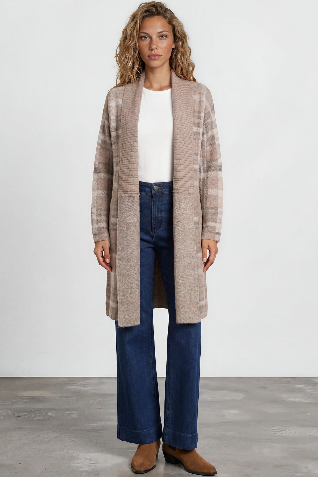 Woman wearing a long beige coat, white top, blue jeans, and brown shoes on a plain background