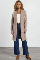 Woman wearing a long beige cardigan, white top, blue jeans, and brown shoes on a plain background