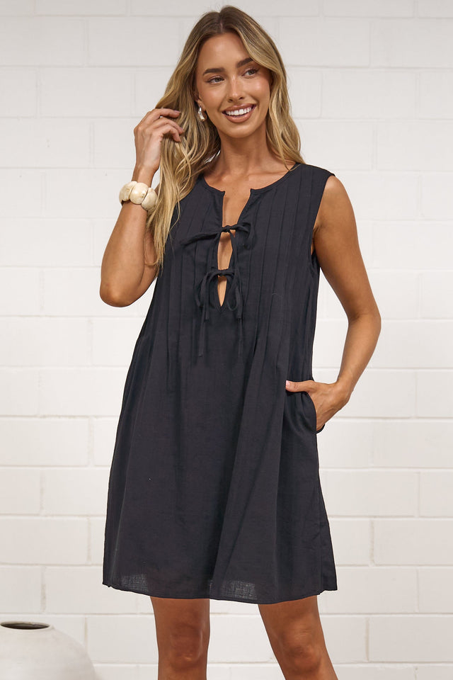 A woman wearing a black sleeveless mini dress with a tie-front neckline and relaxed shift silhouette, standing in front of a white painted brick wall.