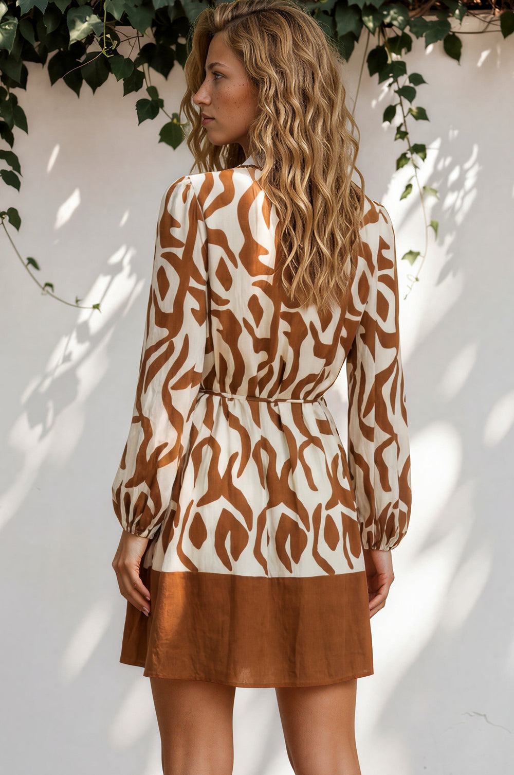 Woman wearing a patterned top and brown skirt standing against a white wall with greenery.