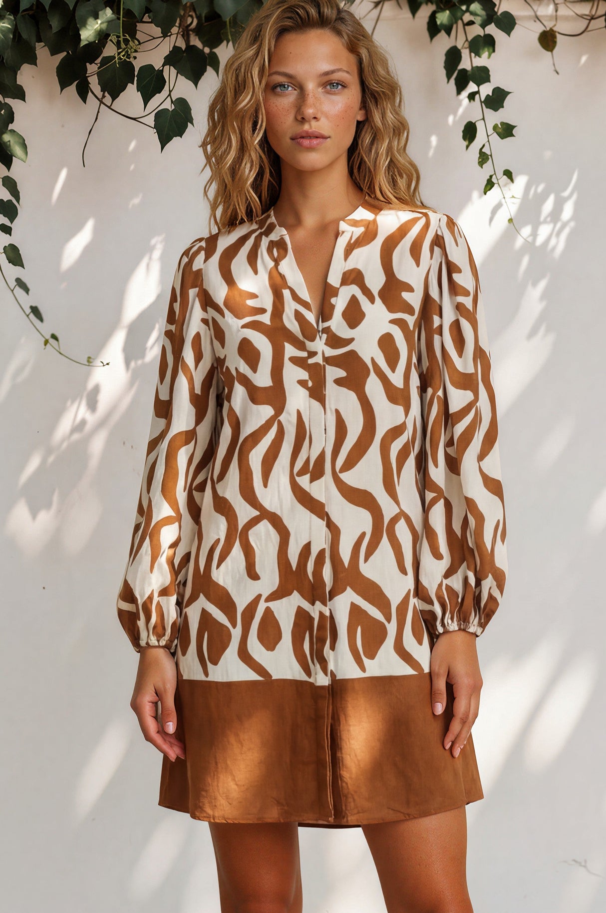 Woman wearing a patterned top and brown skirt standing against a white wall with greenery.